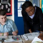 Tutor reading textbook next to male student revising