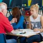 Male tutor in red t-shirt explaining topic to female student in white vest at desk with workbooks