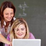 Female tutor helping female student with studies on laptop with equations on a blackboard in the background