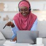 Student with a headscarf studying on a laptop at home on a sofa